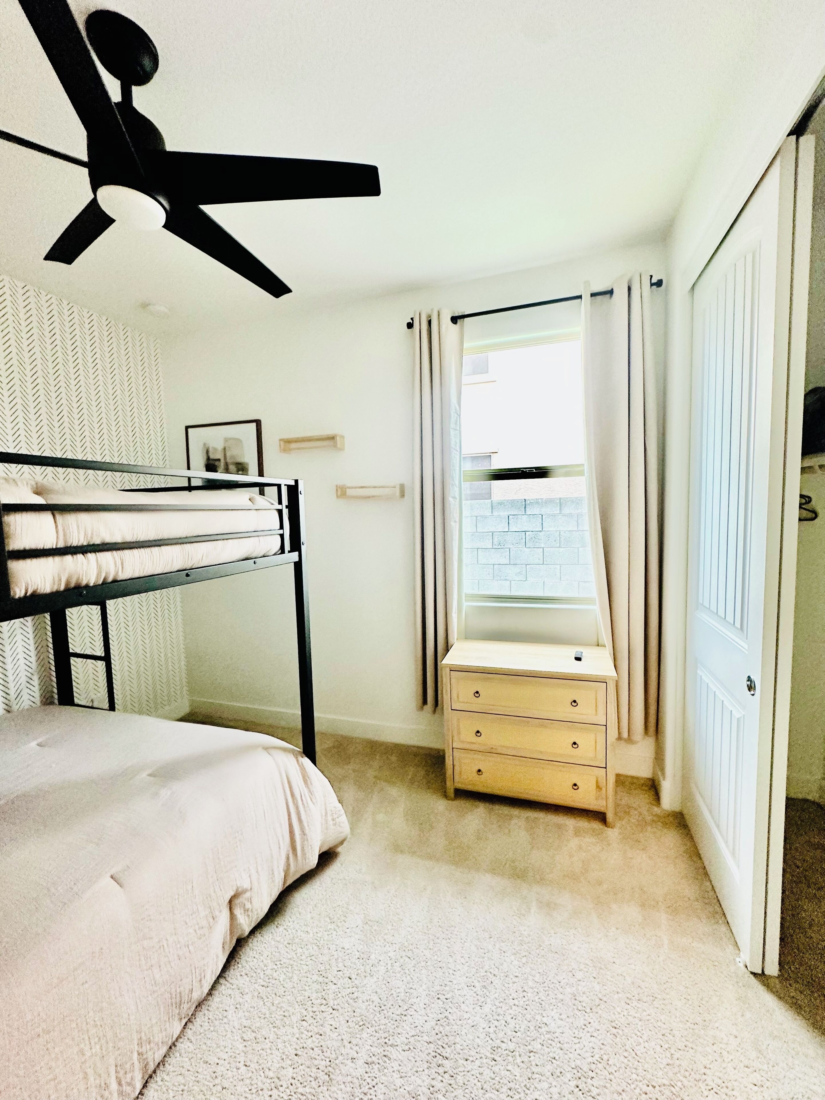 Une chambre avec un lit superposé, un ventilateur de plafond, une petite table de chevet avec des tiroirs, une fenêtre avec des rideaux et une porte donnant sur une autre pièce.