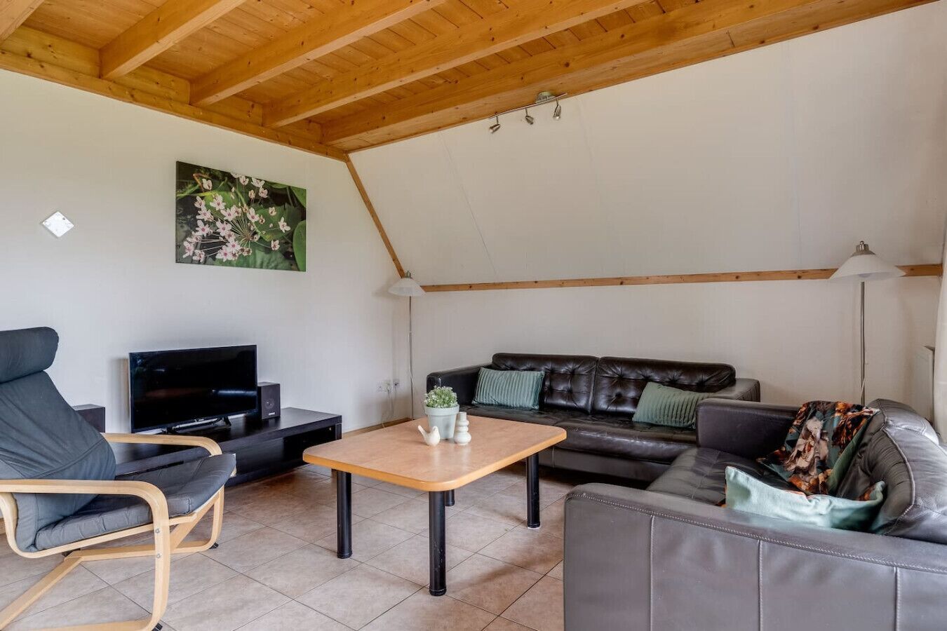 A cozy living room with a wooden ceiling, a black leather sofa, a grey armchair, a wooden coffee table, and a flat-screen TV.
