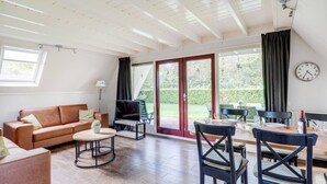 Un vaste espace de vie comprenant un canapé, un téléviseur et une table à manger avec des chaises. Une grande fenêtre, ornée de rideaux, et une horloge murale complètent la pièce.
