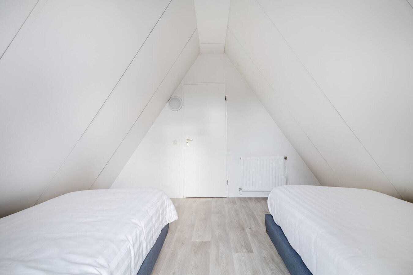 A small, white, attic-style bedroom with two single beds, a door, and a radiator.