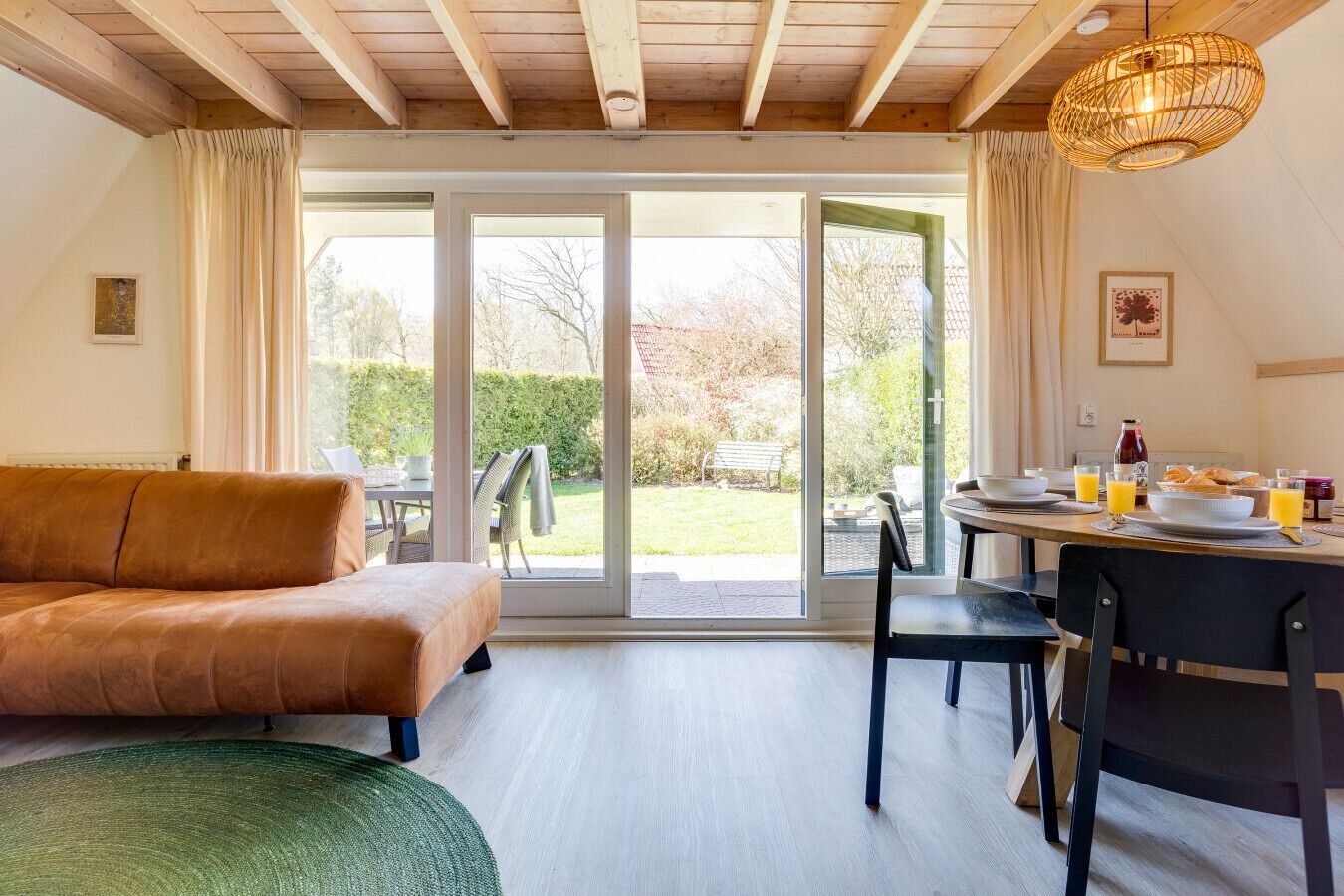 A cozy room with a brown leather sofa, a dining table with breakfast items, and a view of a garden through large sliding glass doors.