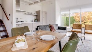 A dining area with a wooden table set for two, a sofa, and a view of a garden through large windows.