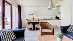 Eine moderne Küche mit weißen Schränken, ein Essbereich mit Korbstühlen und einem Tisch sowie ein Wohnbereich mit einer Couch und einem Couchtisch.