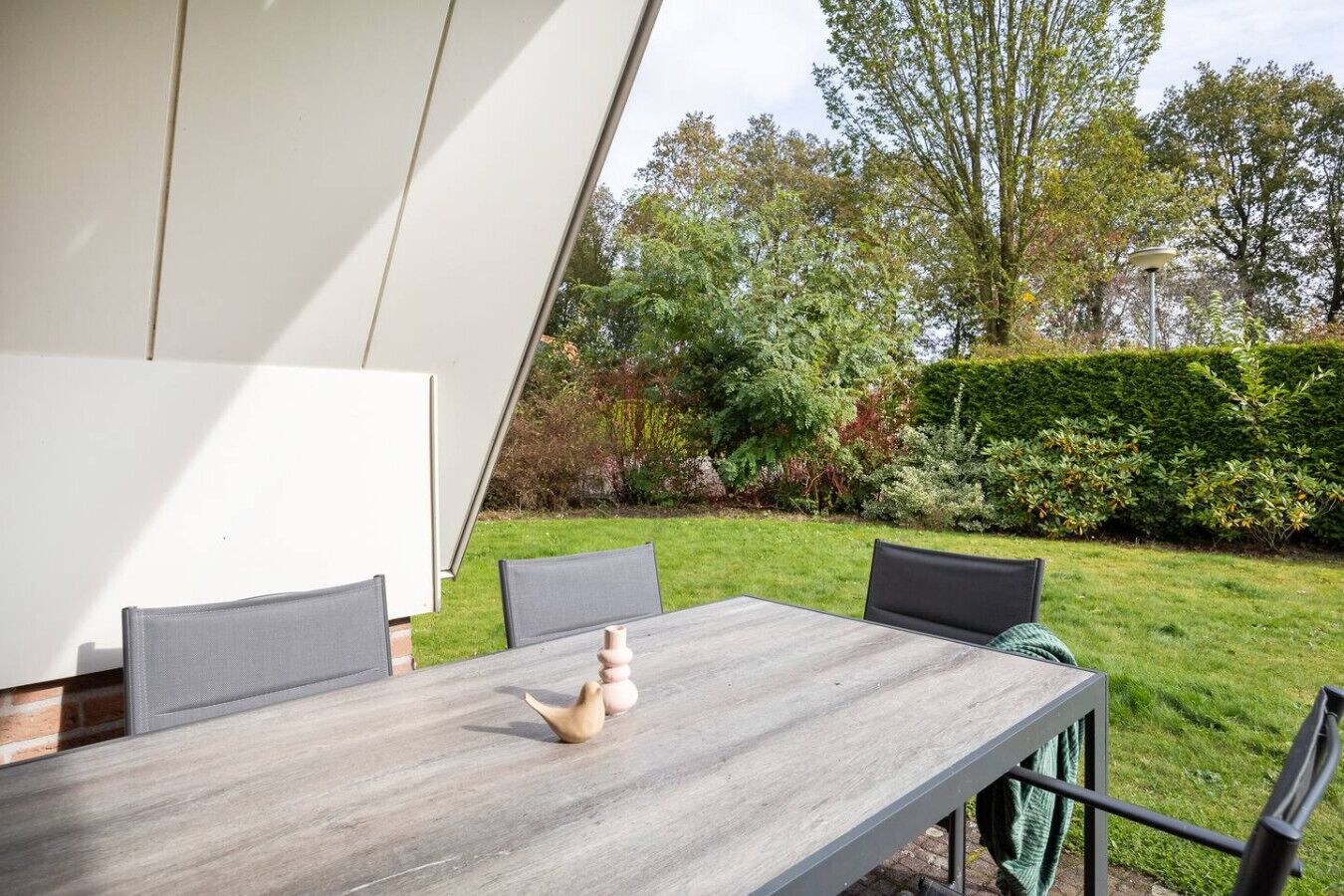 A modern outdoor dining area with a wooden table and chairs, surrounded by a well-maintained garden and a hedge.