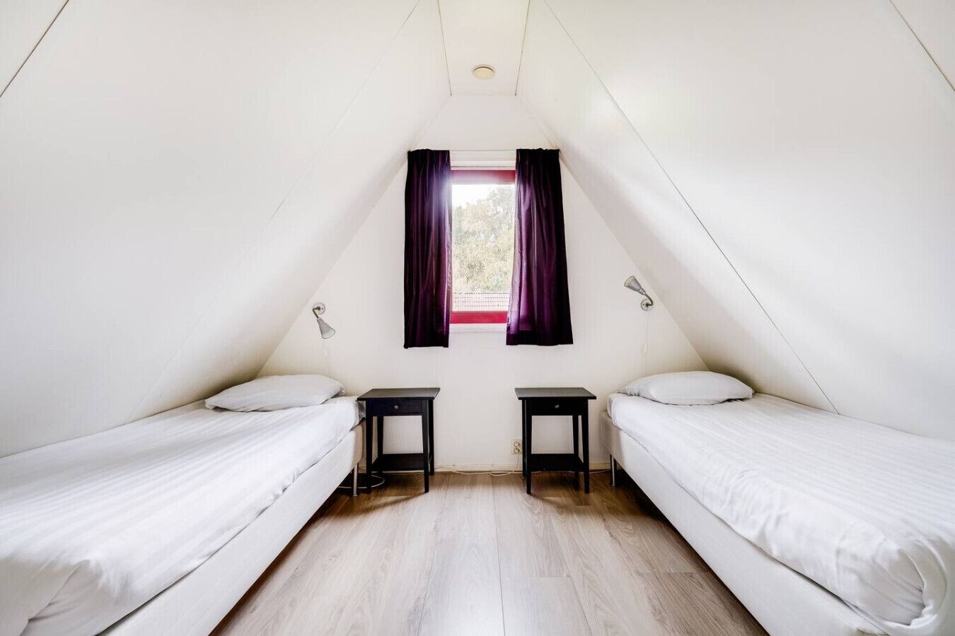 Un pequeño dormitorio en ático, con forma de triángulo, con dos camas individuales, una ventana con cortinas moradas y suelo de madera.