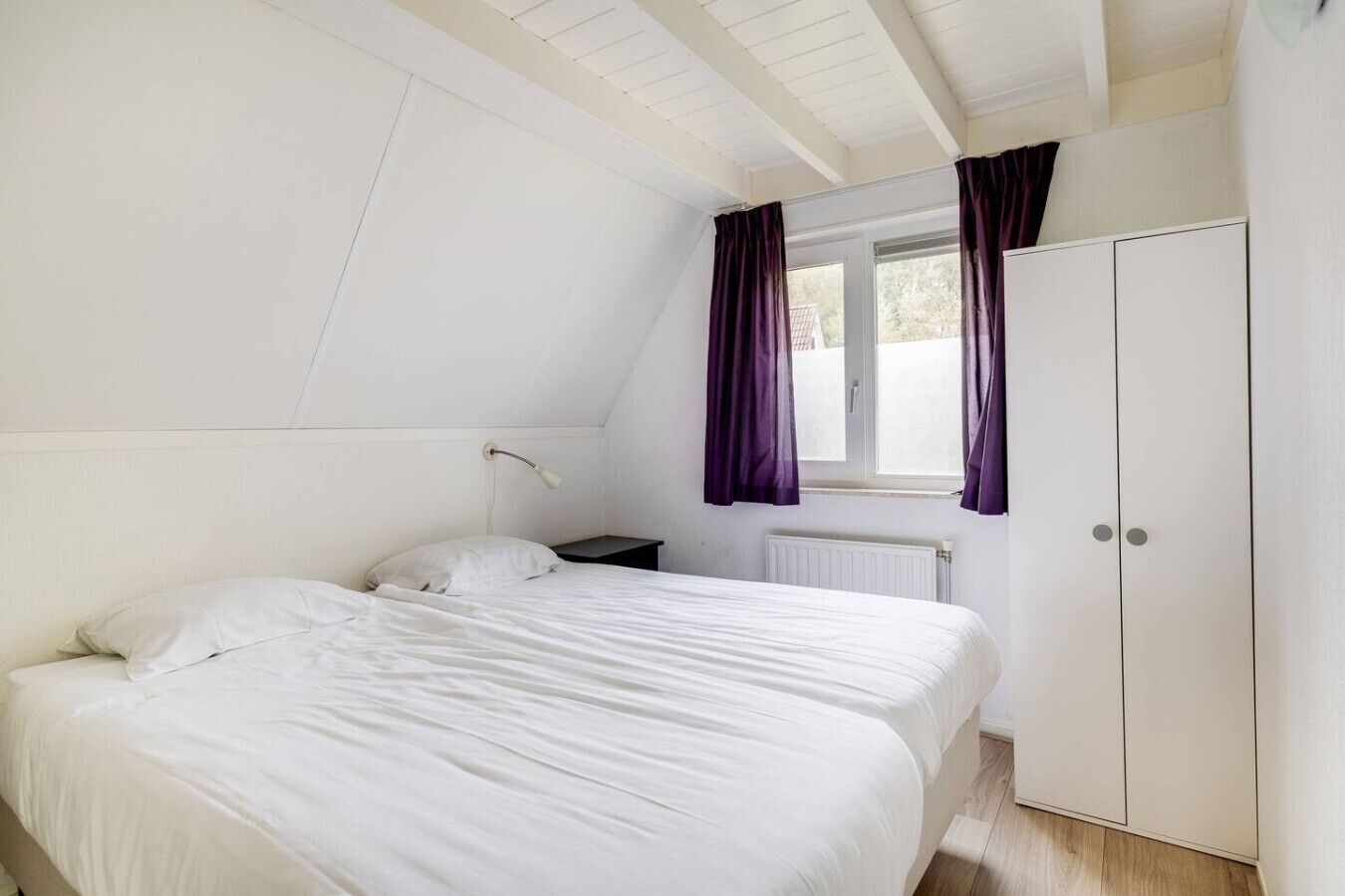 A bedroom with a bed, a nightstand, a window with curtains, and a wardrobe.