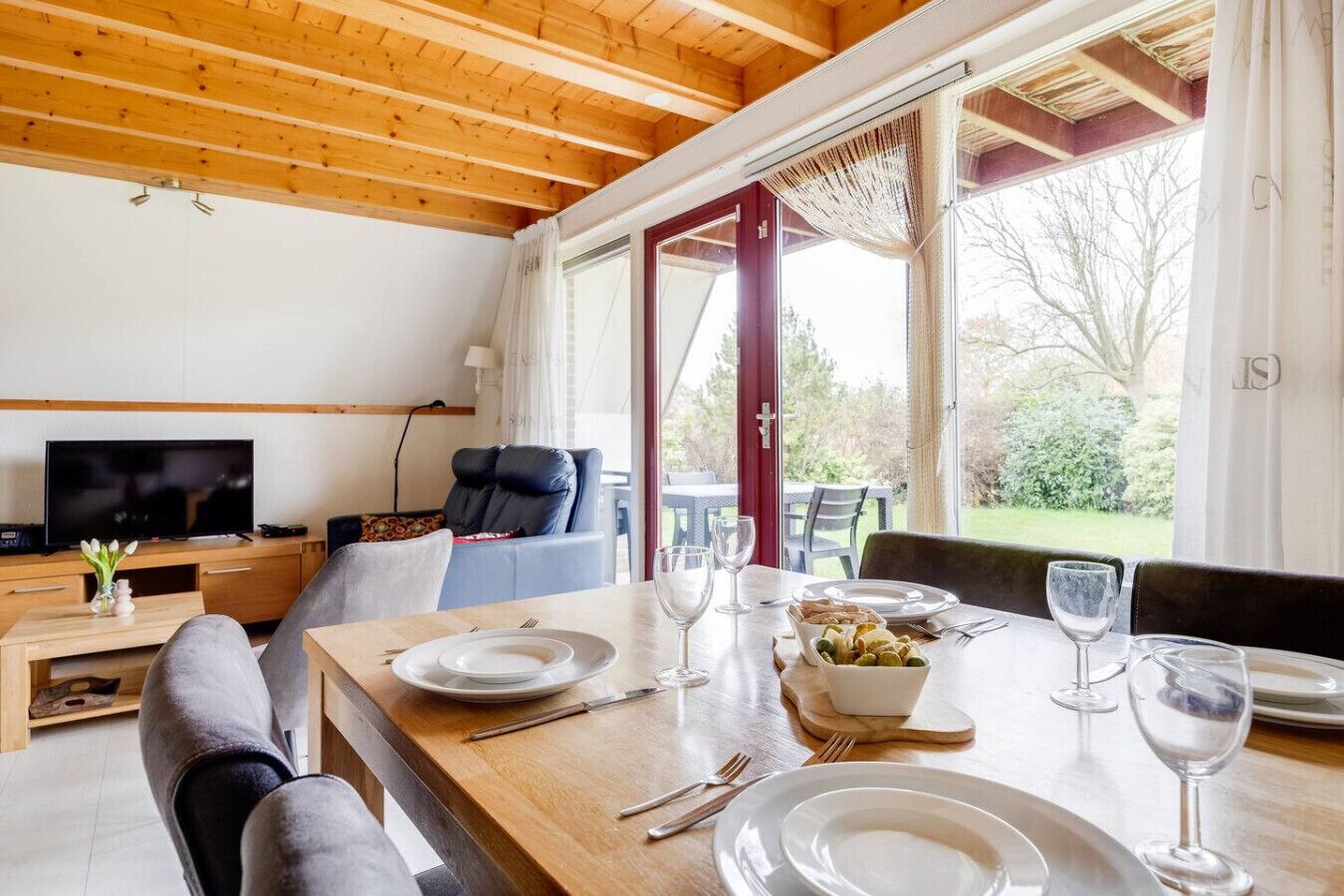 Een eethoek met een houten tafel voor twee personen, een televisie op de achtergrond en uitzicht op het landschap door grote ramen.