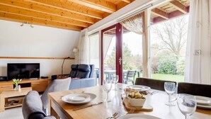 A dining area with a wooden table set for two, a television in the background, and a view of the outdoors through large windows.