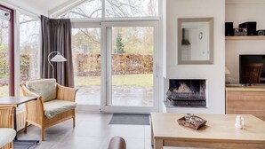 Een moderne woonkamer met een grote schuifpui naar een tuin, een open haard en een televisie op een houten standaard.