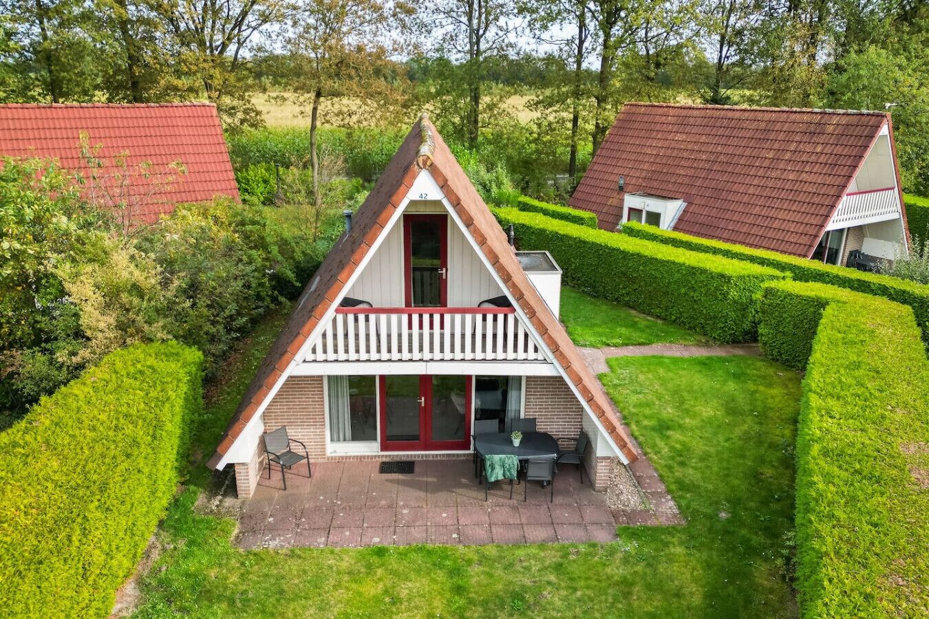 A triangular-roofed house with a balcony, surrounded by well-maintained hedges and a grassy lawn.