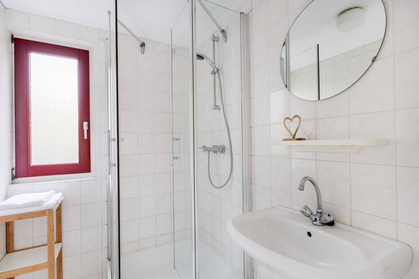 Een moderne badkamer met een douchecabine van glas, een witte wastafel, een ronde spiegel en een raam met rode omlijsting.