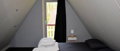 A small, triangular room with a bed, a nightstand with a lamp, and a window with a view of a red-roofed building.