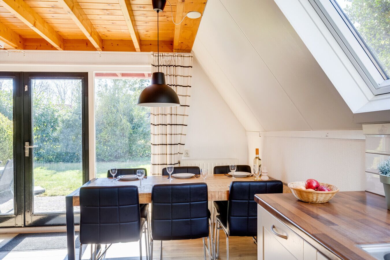 A modern kitchen with a dining area, featuring a wooden ceiling, large windows, and a view of the outdoors.