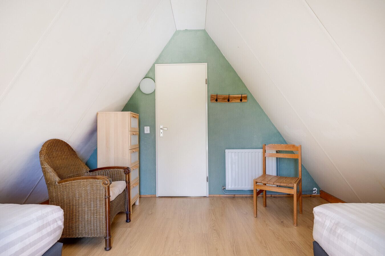 Une petite pièce mansardée triangulaire, avec une porte, deux fauteuils en osier, une chaise en bois et un radiateur.