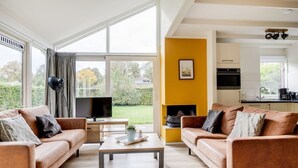 Ein modernes Wohnzimmer mit zwei Sofas, einem Couchtisch und einem Fernseher. Ein Kamin ist zu sehen, und durch eine offene Tür gelangt der Blick in eine Küche.