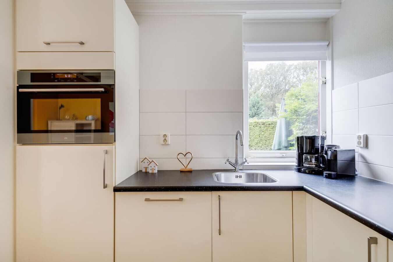 Eine moderne Küche mit weißen Schränken, einer schwarzen Arbeitsplatte, einem eingebauten Backofen, einem Fenster mit Blick auf Grünflächen und einer Kaffeemaschine.