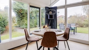 Un comedor con una mesa redonda, cuatro sillas, una araña y ventanas amplias que ofrecen vistas a un jardín y árboles.