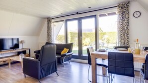 Een moderne woonkamer met een flatscreen televisie, een bank, een eettafel met stoelen en een groot raam met uitzicht op de omgeving.