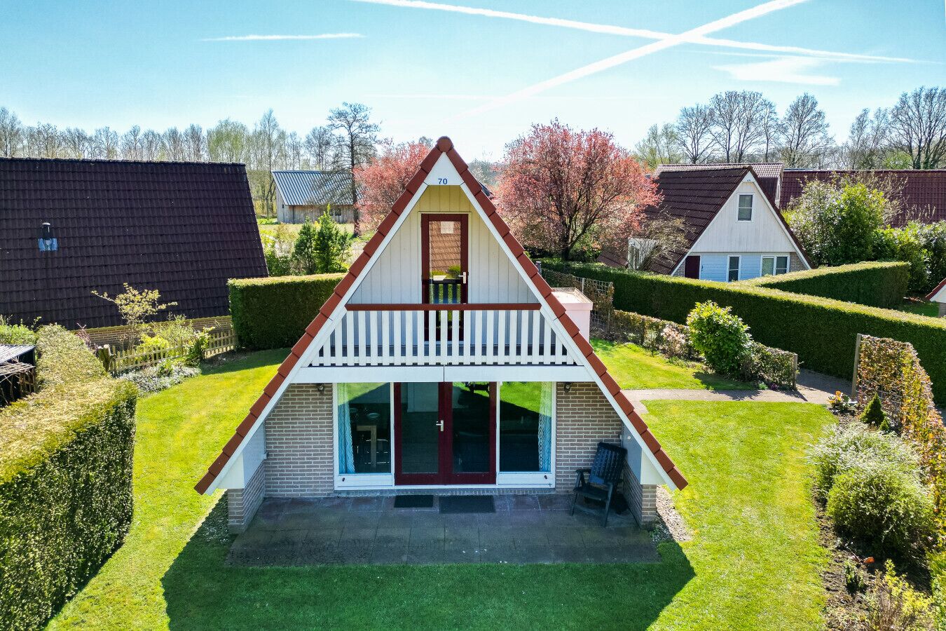 Een driehoekig huis met een balkon, omgeven door verzorgde tuinen en omliggende woningen.