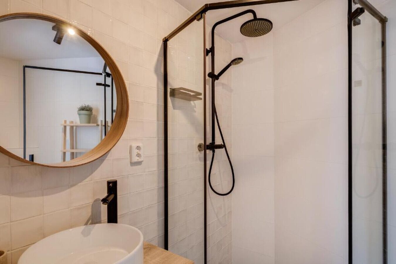 Une salle de bain moderne avec un mur recouvert de carreaux blancs, un miroir rond, un lavabo blanc et une douche noire dotée d’un pommeau de douche à main.