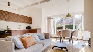 Ein modernes Wohnzimmer mit einer Couch, einem Couchtisch und einem Essbereich mit Stühlen. Im Hintergrund ist eine Küche mit Fischgrätenmuster-Spritzschutz erkennbar.