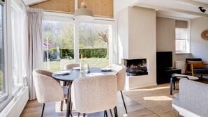 Ein modernes Wohnzimmer mit Kamin, einem Essbereich mit rundem Tisch und Stühlen sowie einem großen Fenster mit Blick ins Freie.