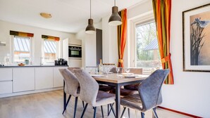 A modern kitchen with white cabinets, a dining area with a wooden table and grey upholstered chairs, and a large window with colorful curtains. - Cuckoo I Modern Home with Private Garden (Zwiggelte)