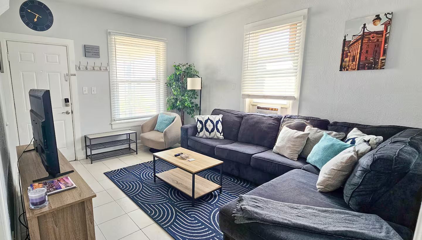 A living room with a sofa, armchair, coffee table, and a TV. There are two windows with blinds, a plant, and a wall clock.
