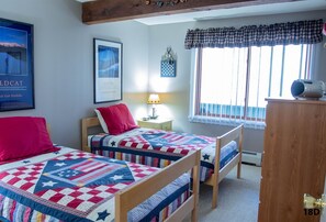 A bedroom with two single beds, a nightstand with a lamp, a window with curtains, and a framed picture on the wall. - Official Seasons RMC Condo 18D - 2 bedrooms - sleeps 4 - No Fees (Bartlett)
