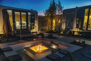 A modern outdoor living area with a fire pit, seating, and a glass-enclosed bedroom.