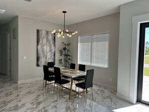 A dining area with a marble floor, a chandelier, black chairs, a white table, and a large window with blinds. - Upscale Getaway (Rotonda West)