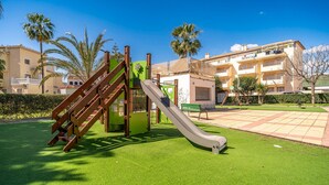 Une aire de jeux avec un toboggan et une structure d’escalade, entourée d’une pelouse bien entretenue et d’un bâtiment en arrière-plan.