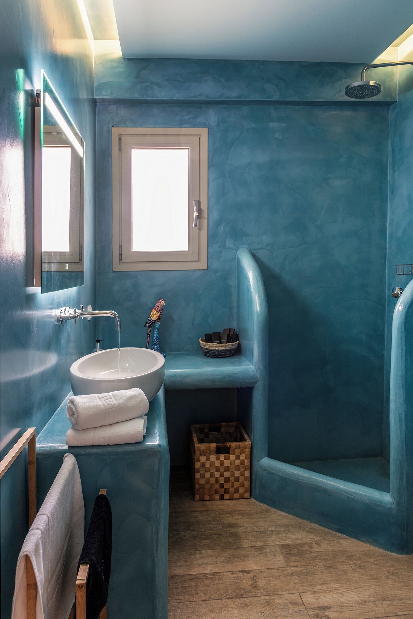 A bathroom with a blue-tinted wall, a white sink, a mirror, a window, and a shower area.