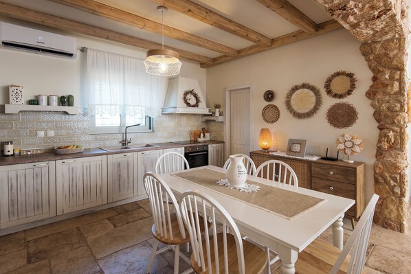 Una cocina con armarios blancos, pared de piedra y comedor con mesa y sillas.