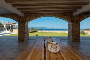 Un espacio con mesas y sillas al aire libre, con arcos de piedra, vista al mar y un edificio de piedra al fondo.