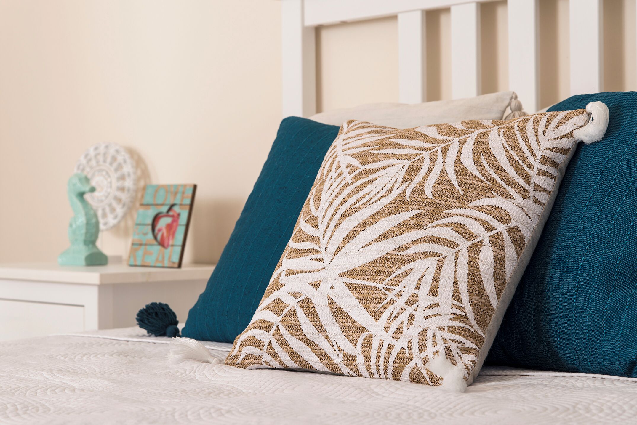 Un lit bien fait, recouvert d’une couette blanche, agrémenté de coussins turquoise et d’un coussin décoratif orné d’un motif de feuilles.