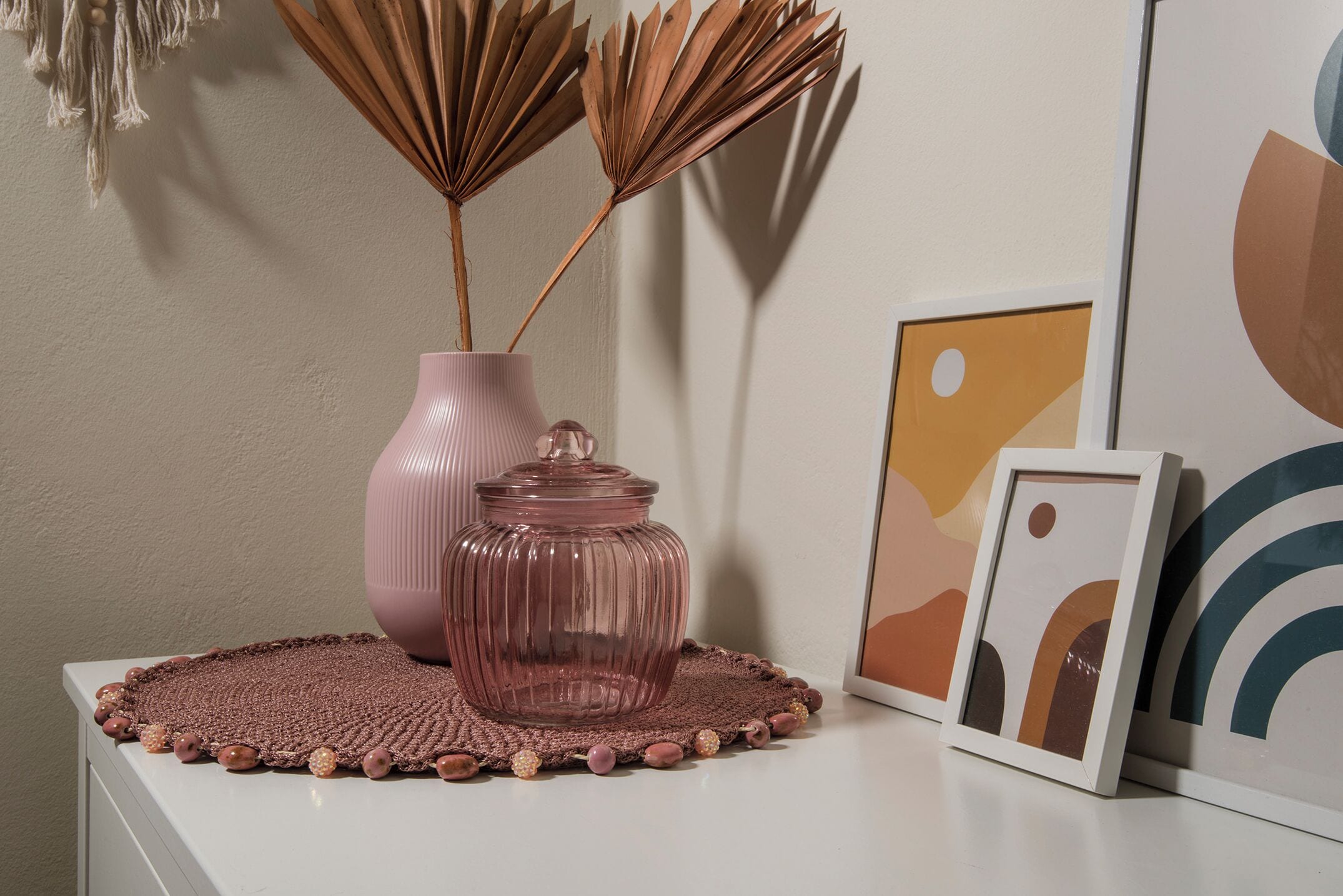 A decorative arrangement on a white surface including a pink vase, a clear glass jar, and a woven placemat.