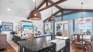 Una cocina con una isla grande, vigas de madera, un comedor con mesa y sillas, y vista al océano.