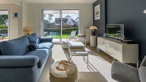 A modern living room with a sofa, a coffee table, a TV, and a sliding glass door leading to a garden. - Village de Légenèse by Interhome (Carnac)