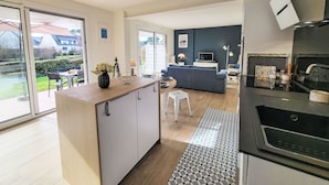 A modern kitchen with a central island, a dining area with a table and chairs, a living room with a sofa and a TV, and a view of a garden through large windows. - Village de Légenèse by Interhome (Carnac)