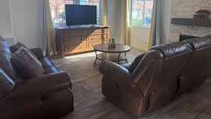 Una sala de estar con sofás de cuero, un mueble de televisión de madera, una mesita redonda con dos piñas y ventanas grandes con cortinas.