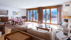 Een ruime woonkamer met een groot raam en uitzicht op de bergen, een gezellige zithoek met banken en een salontafel, en een eethoek met een tafel gedekt voor twee personen.