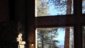 Ein gemütliches Blockhaushaus-Interieur mit Kamin aus Stein, einer gepolsterten Sitzbank mit Blick auf einen Wald und einem Holzbalken.