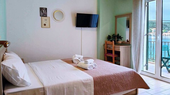 A hotel room with a bed, a flat-screen TV, a desk with a chair, and a balcony with a view of the sea.