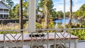 A wooden deck with white chairs and a table, overlooking a lake with trees and a house in the background.