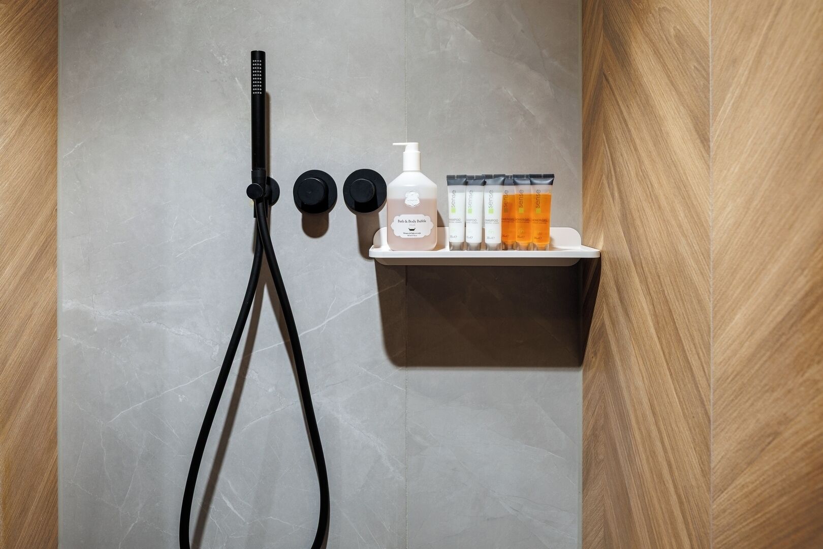 A shower with a handheld showerhead, a wall-mounted control knob, and a shelf with toiletries including a bottle of shampoo and a bottle of body wash.