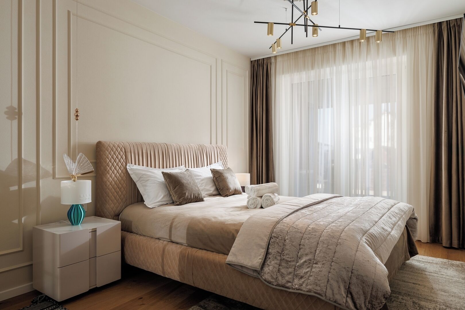 Une chambre à coucher avec un grand lit, une table de chevet avec une lampe et une vue sur l’extérieur à travers des rideaux légers.