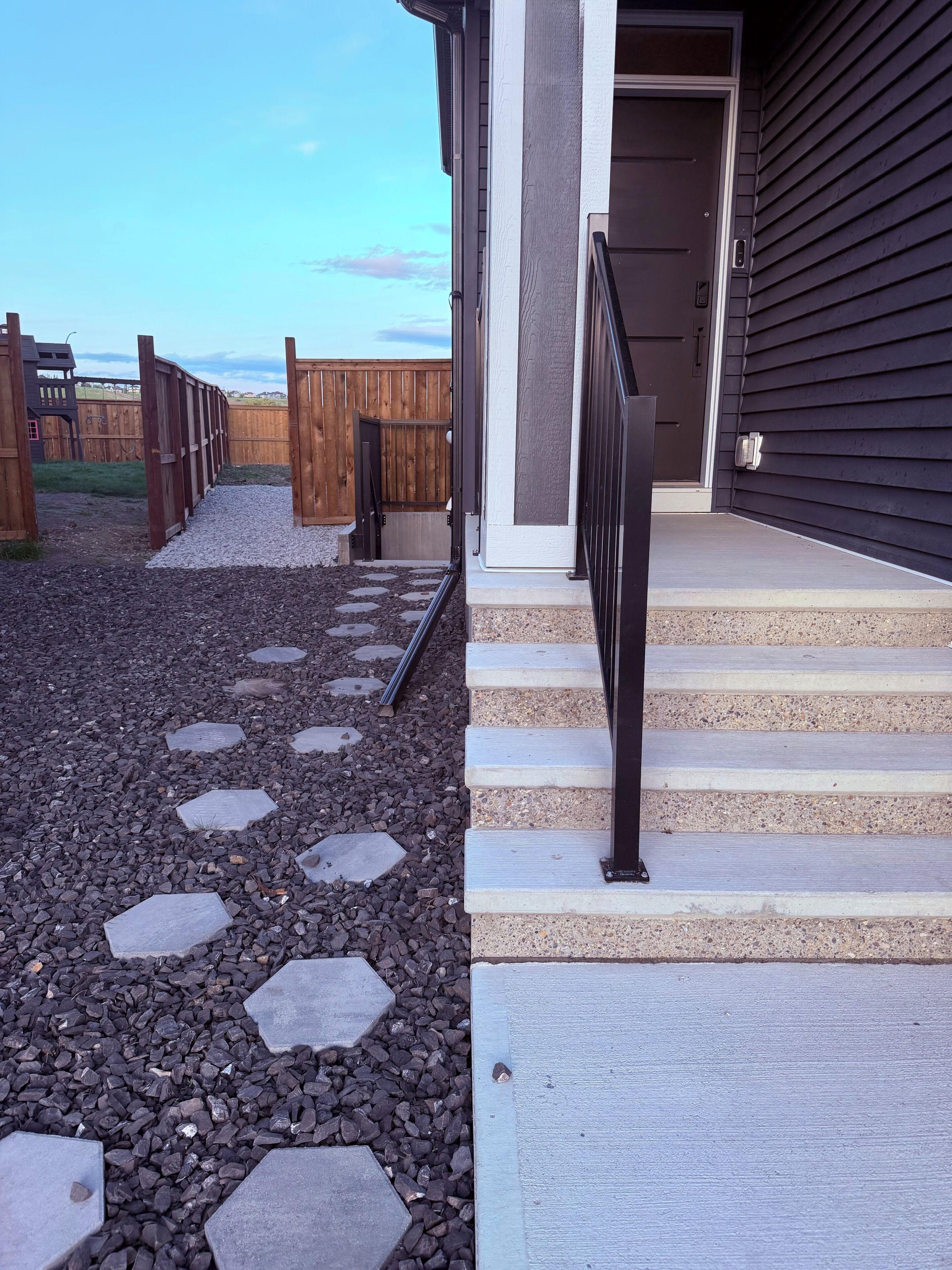 Un chemin menant à une maison entourée d’une clôture en bois, dotée d’une porte sombre et d’un escalier en béton.