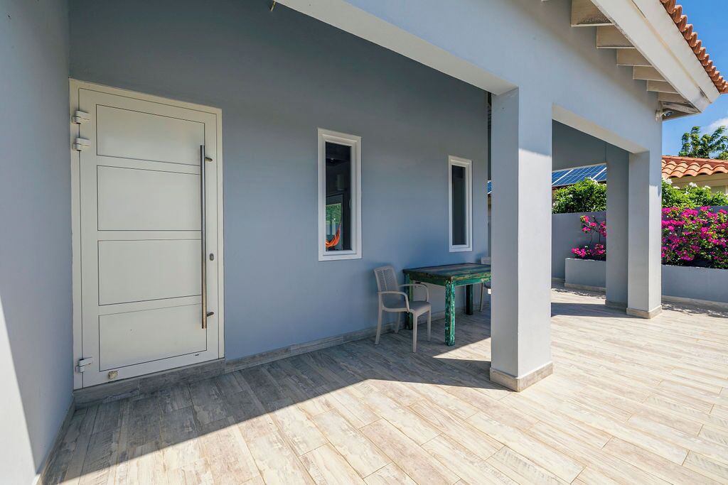 Um espaço coberto ao ar livre com uma mesa, cadeiras, uma porta e janelas.