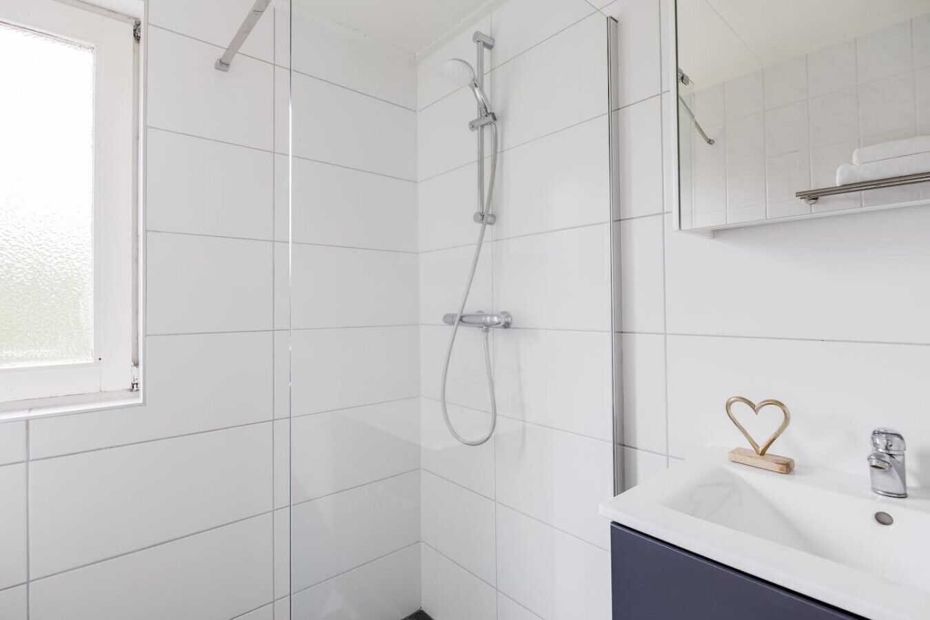 Un baño moderno con azulejos blancos, ducha con cabezal de mano, lavabo con espejo y un estante pequeño con una decoración en forma de corazón.